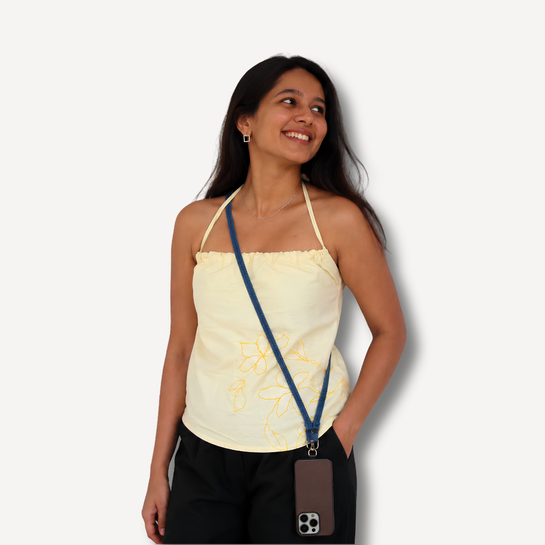 A woman wearing a pale yellow floral print halter top and black pants, carrying a brown phone case on a blue strap around her neck, set against a plain light gray background.