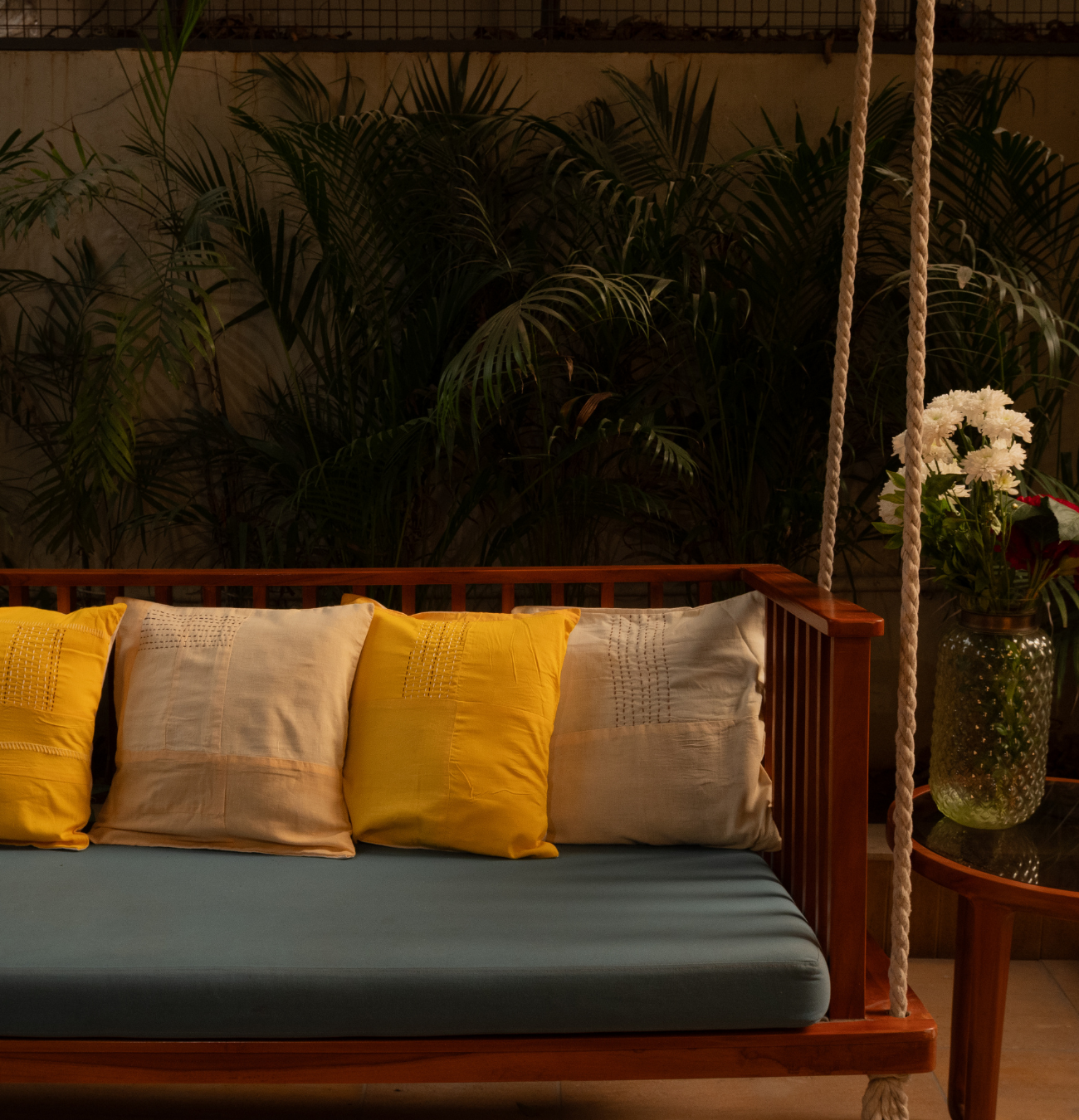 Handcrafted yellow and beige patchwork pillow covers with kantha stitch detailing, styled on a wooden swing sofa with lush green plants in the background - JOD