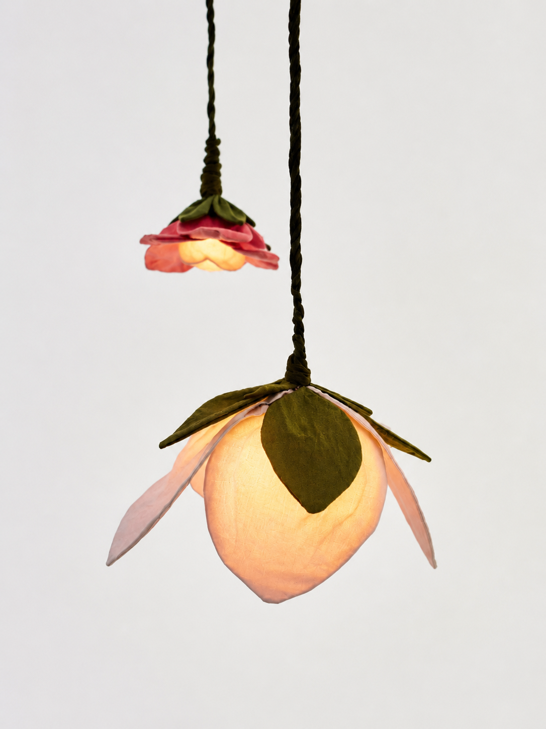 A hanging pendant light shaped like a flower, with a glowing warm white center and pink petals, suspended by a black cord against a plain light gray background.