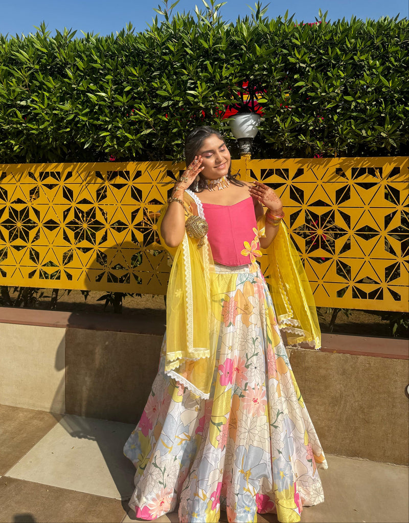 Woman in a colorful traditional outfit standing in front of a yellow decorative wall with greenery.