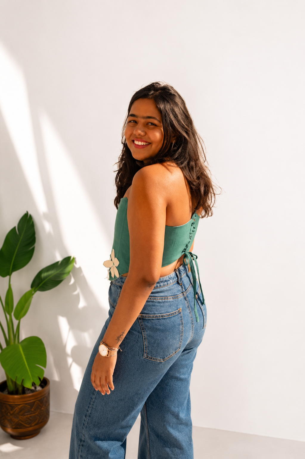 Woman wearing a green top and blue jeans standing next to a plant against a white wall.