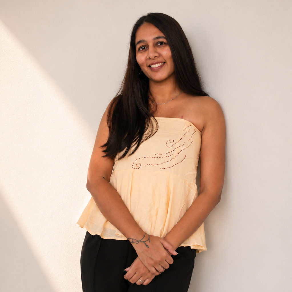 A woman with long dark hair stands against a plain light wall, wearing a strapless pale peach top with small dark embellishments and black pants.