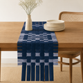 A navy blue woven table runner with a fringe hem is styled on a wooden dining table. The runner features a geometric basketweave pattern in shades of navy, medium blue, and light blue.