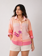A woman modeling a pink and peach striped button-up shirt with purple floral embroidery on the front, paired with light beige shorts, against a plain light gray background.