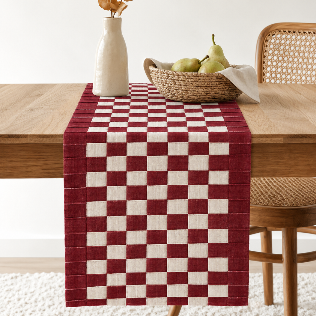 A red and white checkered table runner draped over a wooden dining table, styled with a white vase of dried leaves and a woven basket of pears, with a cane-back chair on the right.
