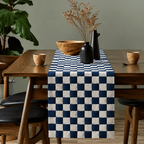 A navy blue and white checkered table runner draped over a wooden dining table, styled with a wooden bowl, dried flowers in black vases, and small wooden cups, set against a muted green wall.