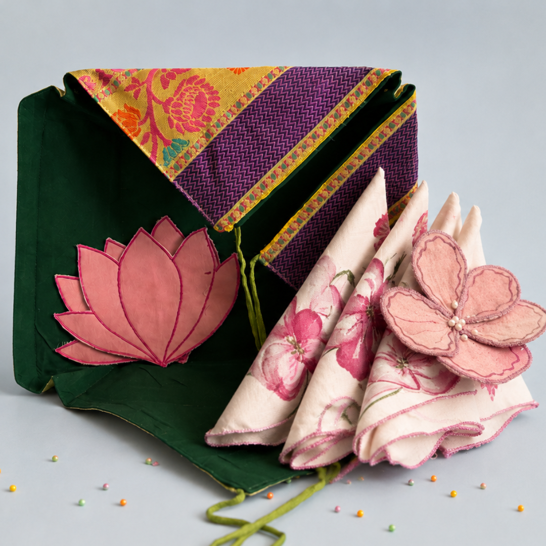 A folded fabric accessory set on a light gray background, featuring a dark green pouch with a pink lotus applique and a purple patterned flap, alongside several cream napkins with pink floral prints and fabric flower embellishments.