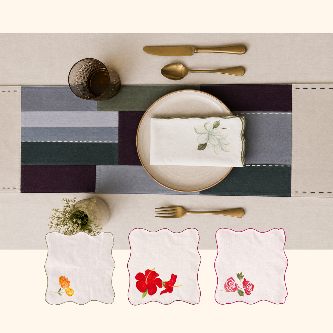 Three square white linen napkins with scalloped edges are displayed below a place setting, each featuring different embroidered floral designs. The napkins are shown against a light beige table background with a striped table runner, gold-toned cutlery, and a small vase of flowers.