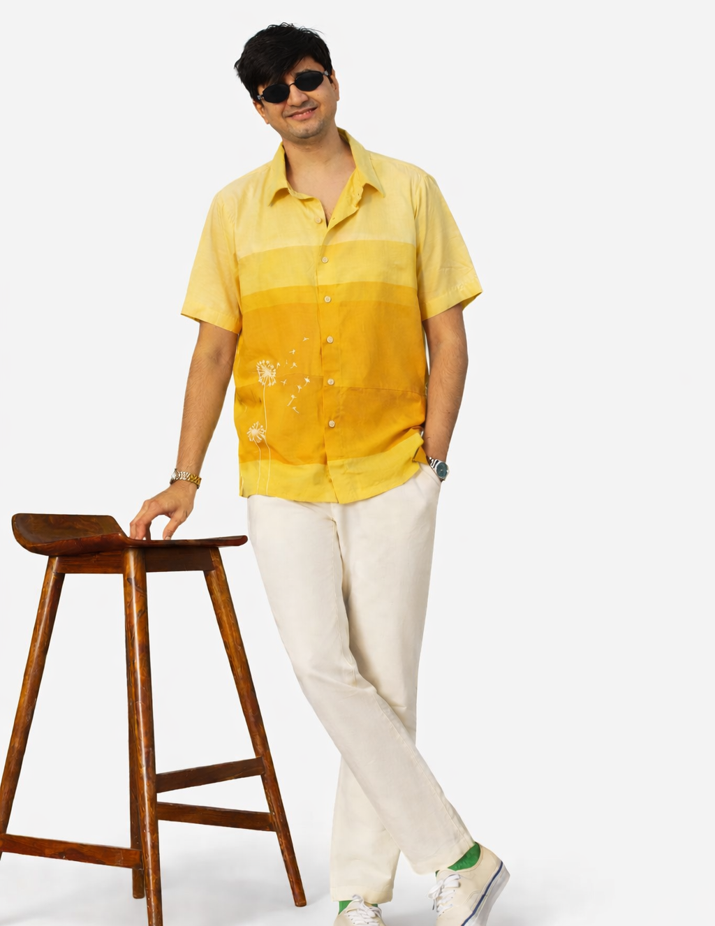 Man wearing a yellow shirt and white pants standing next to a wooden stool on a white background