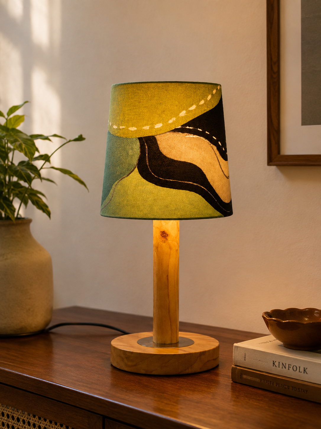 A table lamp with a cylindrical fabric shade featuring an abstract black, olive green, and beige pattern, set on a wooden side table next to a potted plant and books.