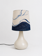 A table lamp with a glossy cream ceramic base and a fabric drum shade featuring a cream upper section with blue and gray wavy lines and a dark navy textured lower section.