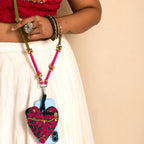 A close-up of a person wearing a red embroidered top and white skirt, carrying a pink and green heart-shaped fabric pouch with a floral pattern over a light blue phone, using a matching crossbody strap.