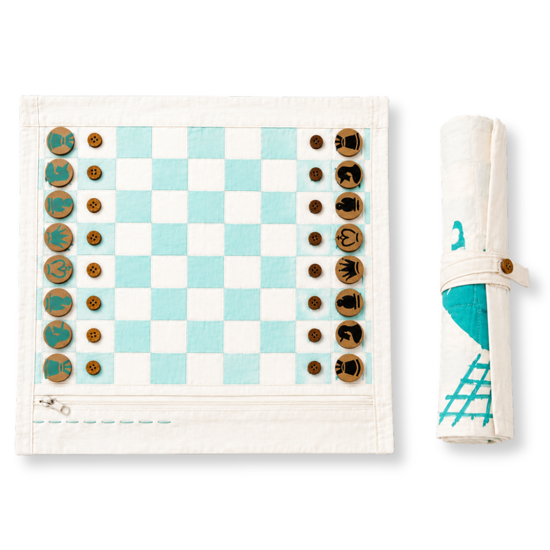 A fabric chess set displayed flat on a white background, next to its rolled-up storage sleeve. The set features a cream-colored canvas board with light blue and white checkered squares, wooden chess pieces with teal and brown details, and a matching rolled-up sleeve with a teal graphic print.