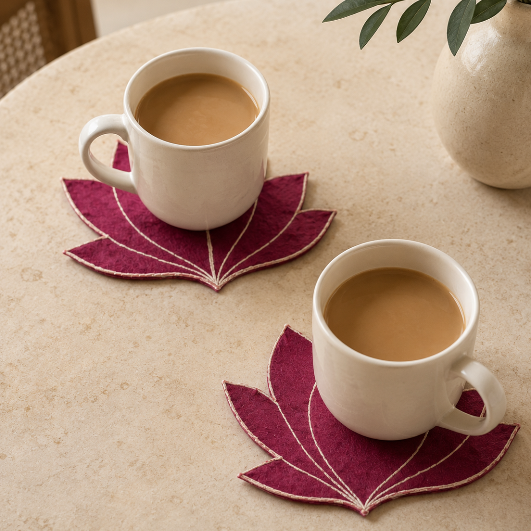 Two white ceramic mugs filled with coffee or tea sit on matching leaf-shaped placemats on a beige tabletop. The COASTERS are deep magenta with white stitched vein details.