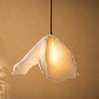 A hanging pendant light with a translucent white fabric flower-shaped shade glowing warm yellow, suspended by a twisted brown cord against a warm beige wall.