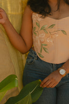 Person wearing a light pink corset top with floral hand painted details, sitting outdoors with greenery.