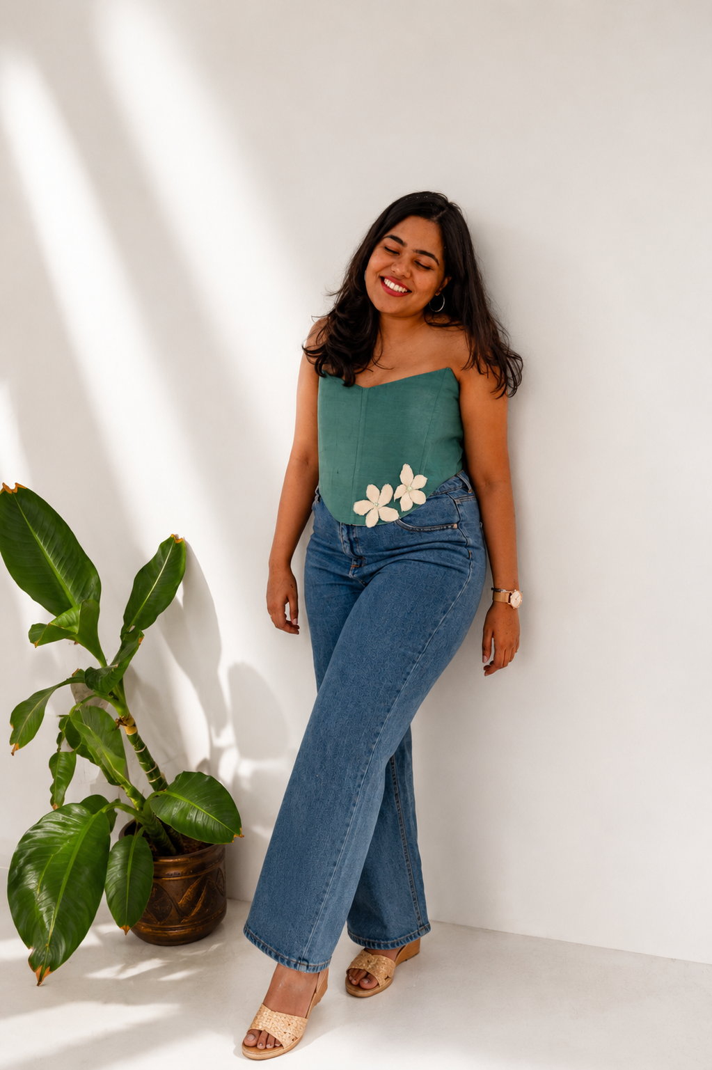 Woman wearing a green top with floral design and blue jeans standing next to a plant against a white wall.