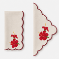 A set of two cream-colored linen napkins with scalloped edges, featuring painted red hibiscus flowers and green leaves, displayed on a light gray background.