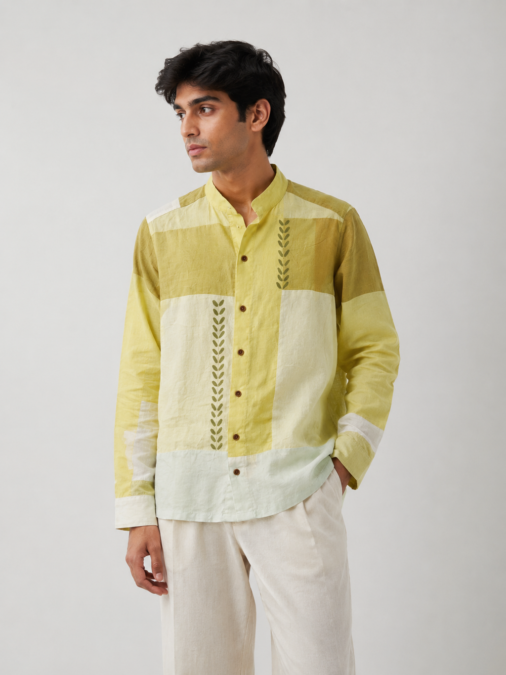 A man wearing a light green and off-white patchwork button-up shirt with embroidered details, paired with off-white trousers, photographed against a neutral light gray background.