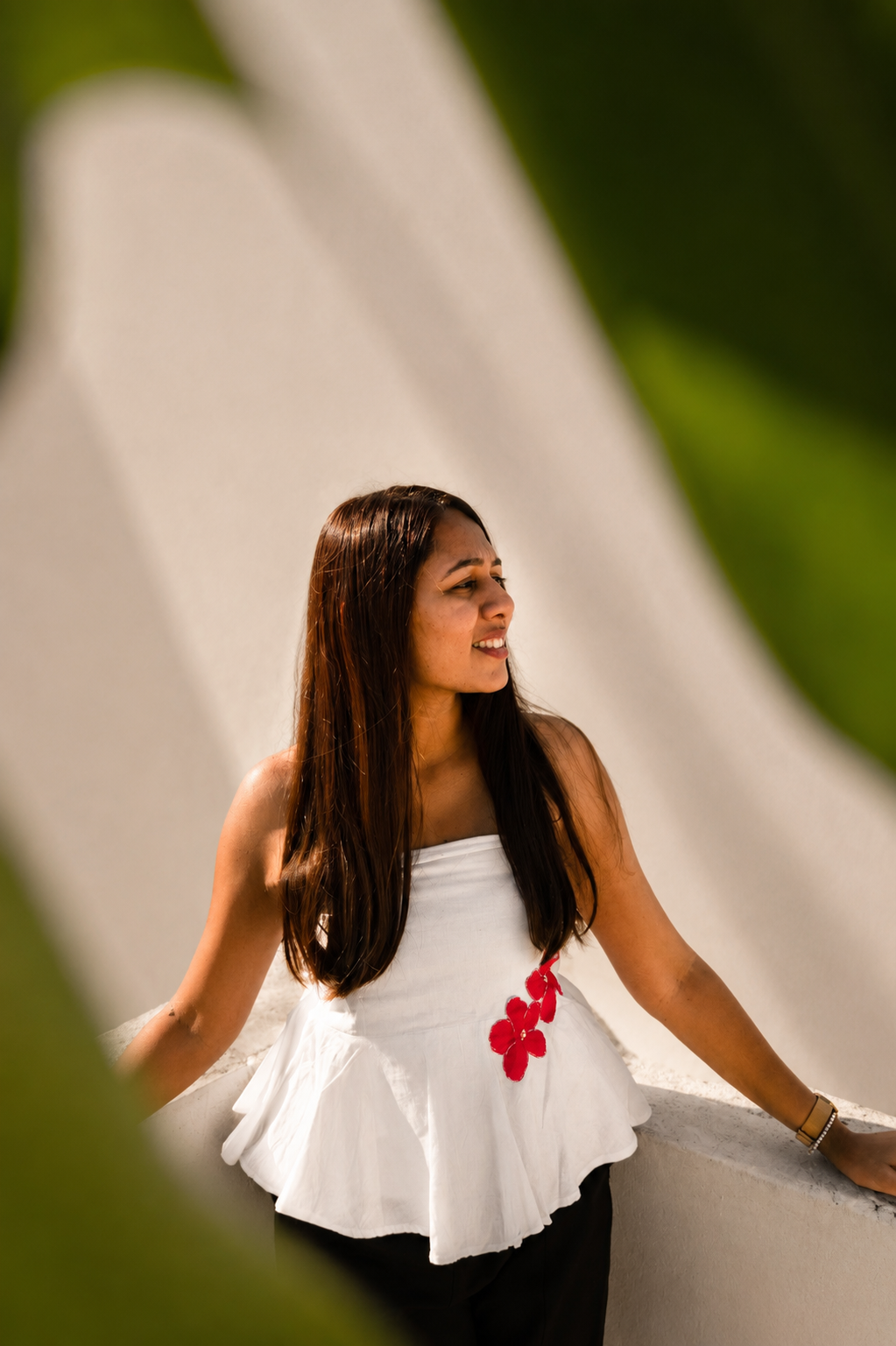 A woman with long dark hair wearing a white strapless ruffled top with red floral accents and black bottoms, leaning against a white wall in a sunny outdoor setting with green leaves framing the shot.