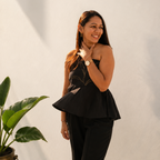 Woman in a black outfit standing against a white wall with a plant to her left.
