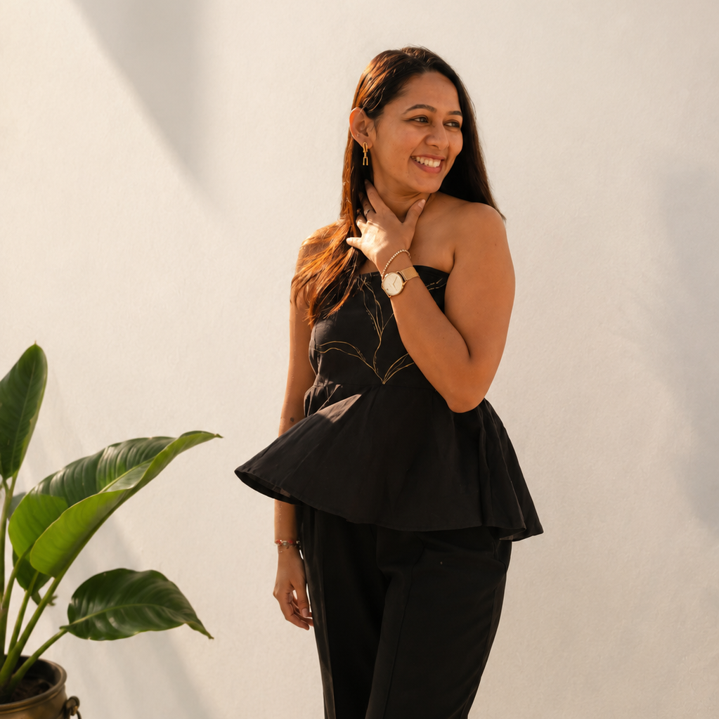 Woman in a black outfit standing against a white wall with a plant to her left.