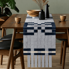 A woven table runner with a geometric pattern in navy blue, light gray, and cream, styled on a wooden dining table with vases and dried flowers.