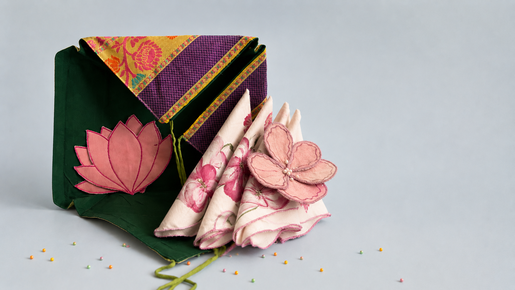 A folded dark green fabric pouch or organizer with a pink lotus applique, shown next to matching rolled napkins and a pink fabric flower on a light gray background.