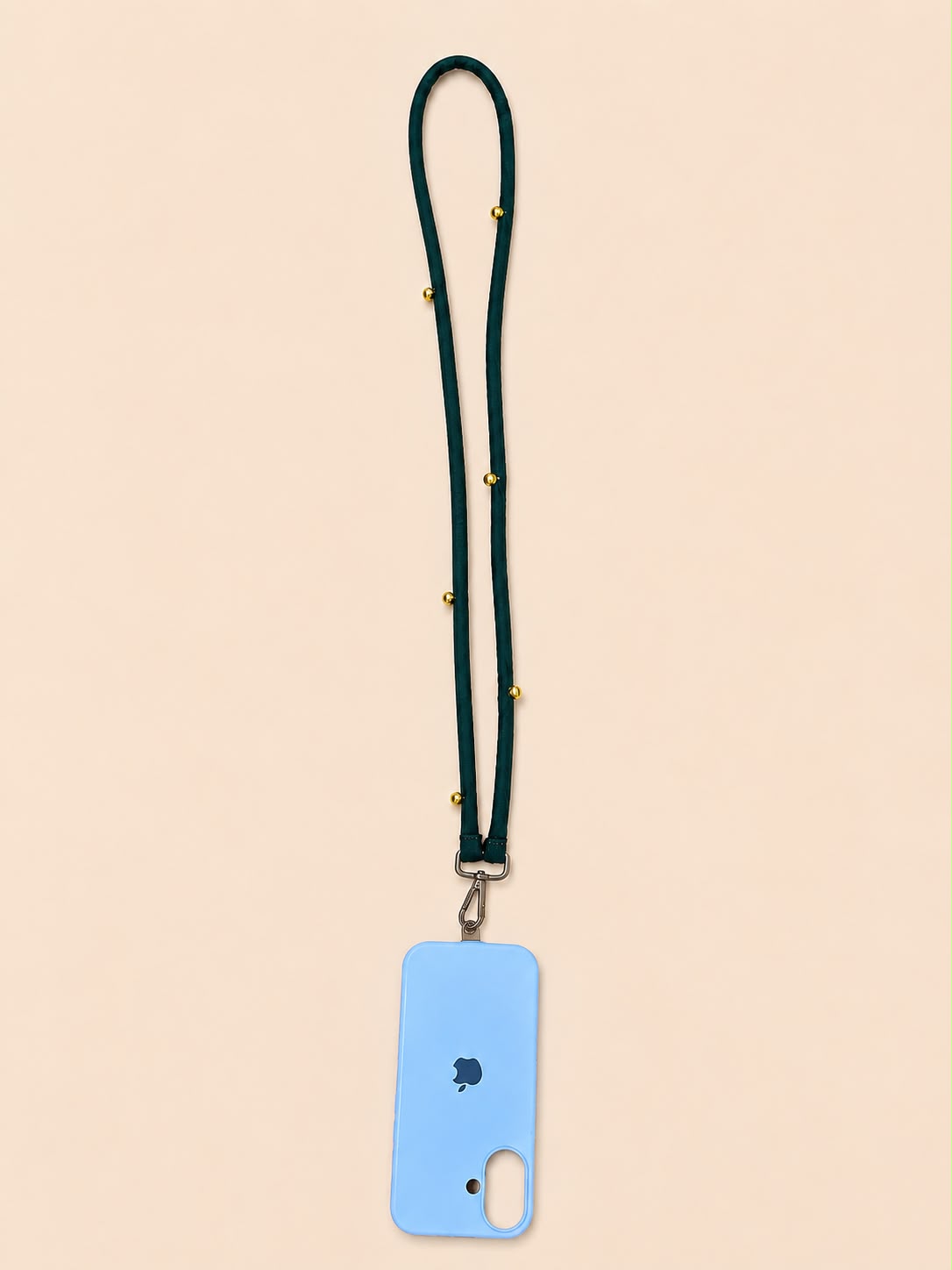 A light blue phone case with a dark green lanyard strap adorned with small gold studs, shown on a solid pale peach background.
