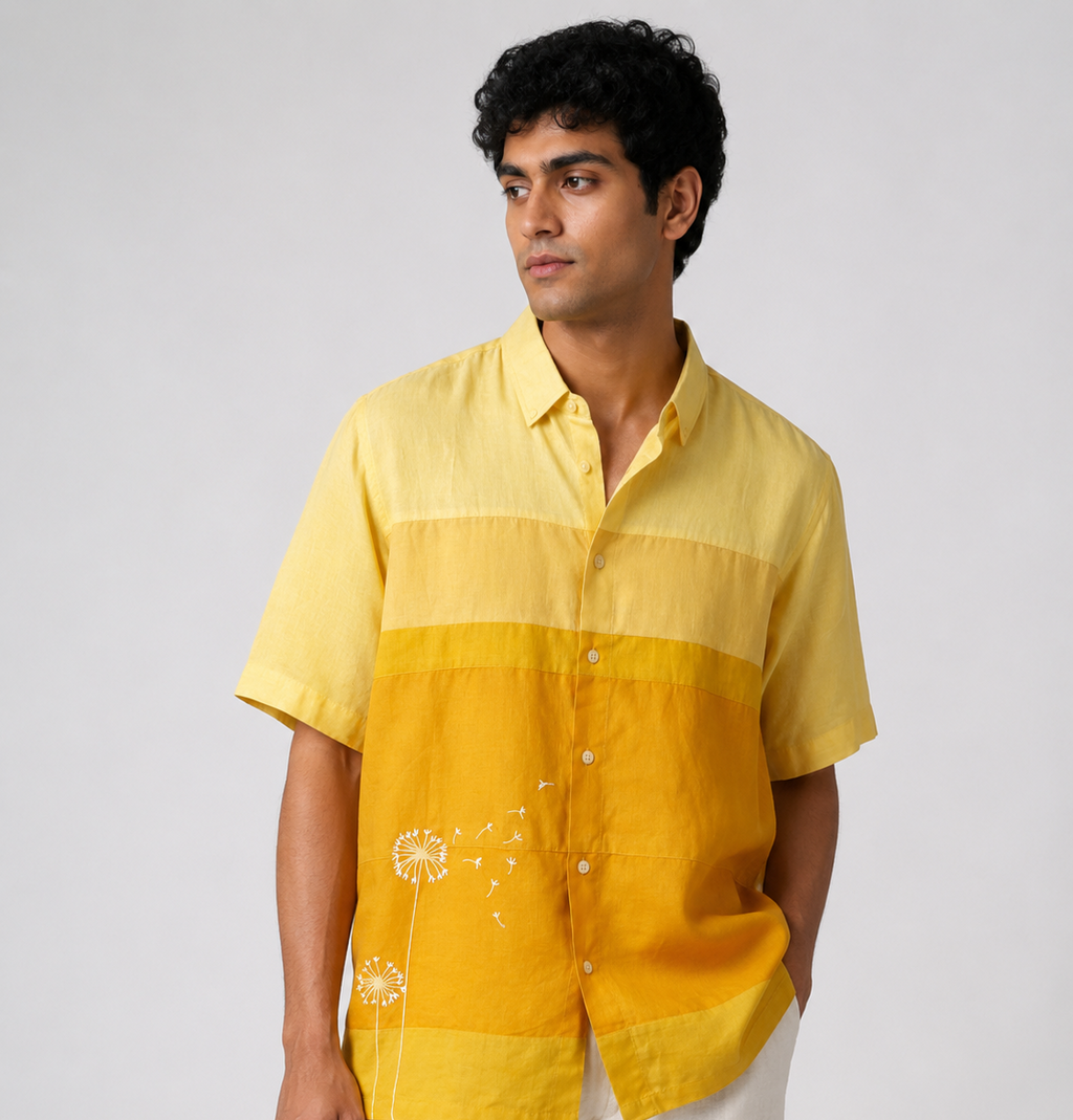 A man wearing a short-sleeve button-up shirt with horizontal yellow and golden-yellow color blocking and white embroidered dandelion details, paired with off-white trousers, against a light gray studio background.