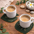 Two white coffee cups on green leaf coasters with a wooden background and decorative elements.
