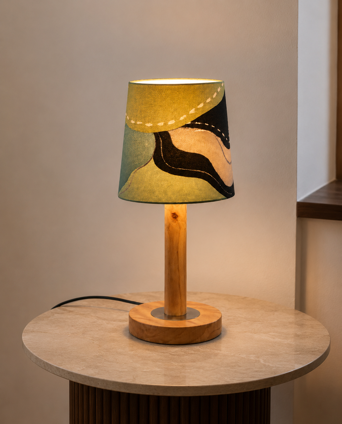 A table lamp with a cylindrical wooden base and a tapered fabric shade featuring an abstract black, beige, and teal pattern, illuminated and placed on a round stone side table.