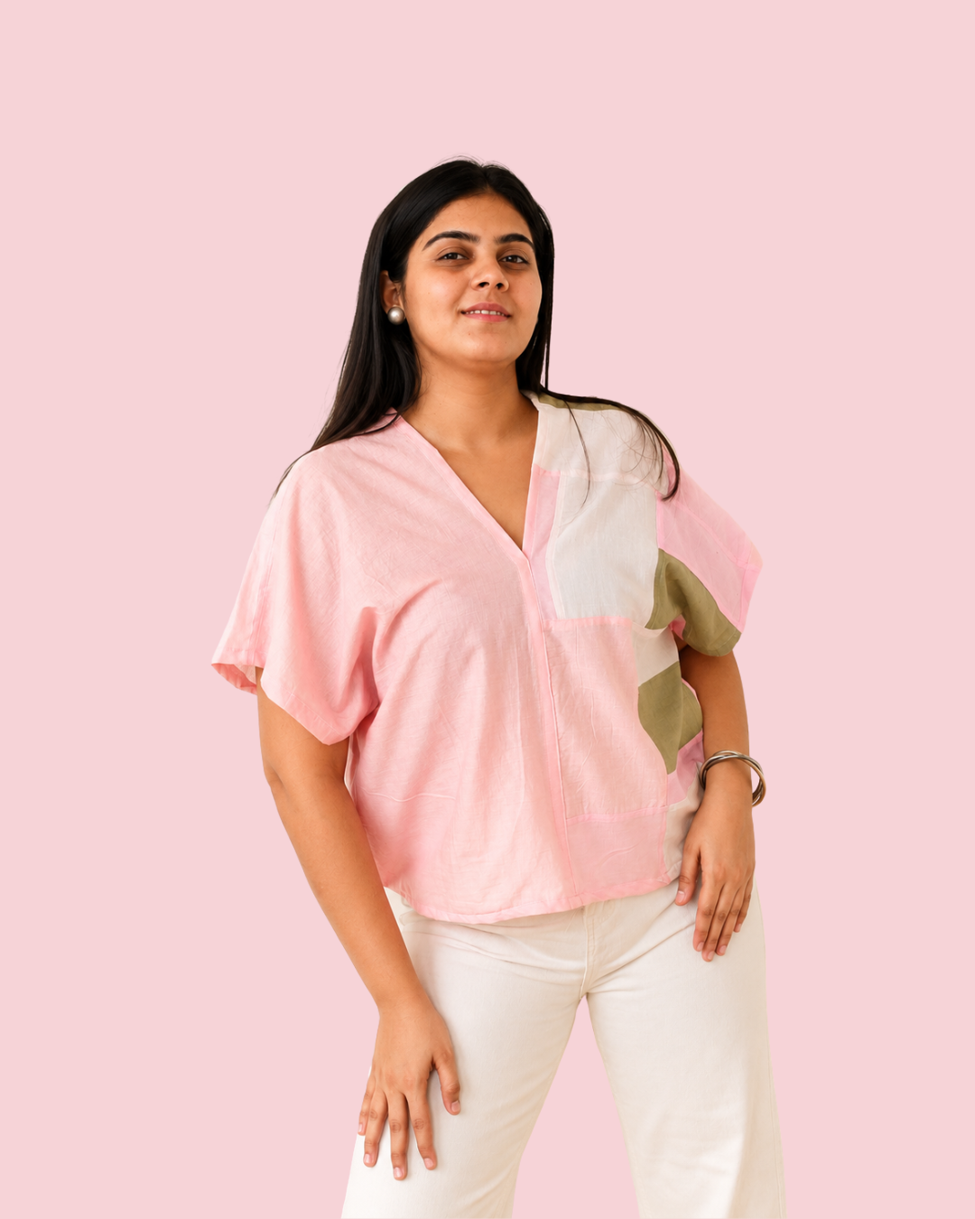 A woman modeling a light pink short-sleeve blouse with white and olive green color-block panels, paired with white pants, set against a solid light pink background.