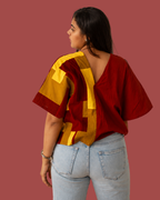 A woman shown from the back wearing a loose-fitting short-sleeve top with a bold geometric block pattern in deep red and mustard yellow, paired with light-wash jeans against a solid muted red background.