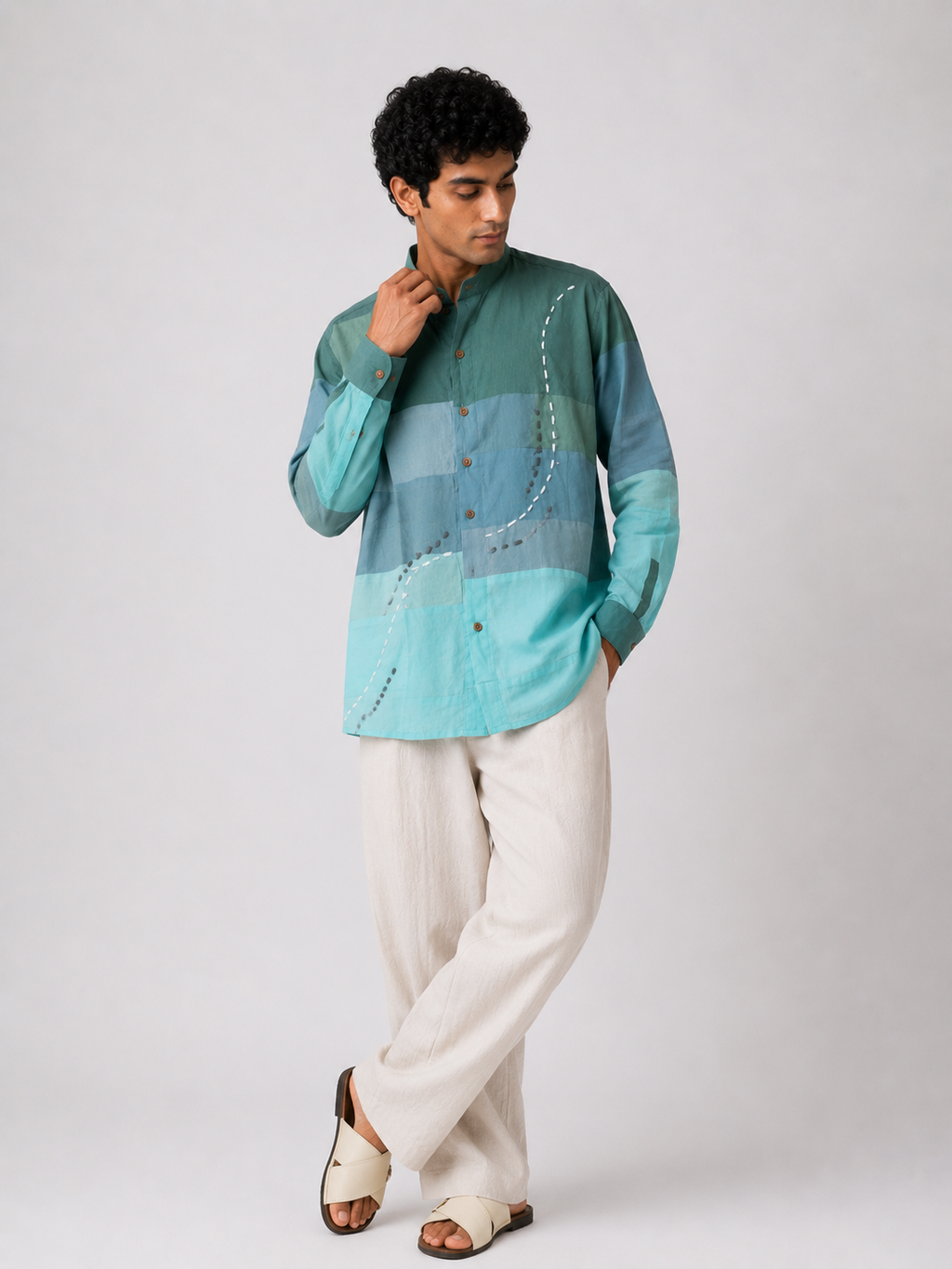 A man modeling a long-sleeve button-up shirt with horizontal stripes in shades of teal, blue, and mint green, paired with off-white trousers and brown sandals, against a light gray studio background.