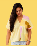 A woman wearing a pastel patchwork short-sleeve blouse with yellow, pink, and orange sections, paired with light blue jeans, set against a solid yellow background.