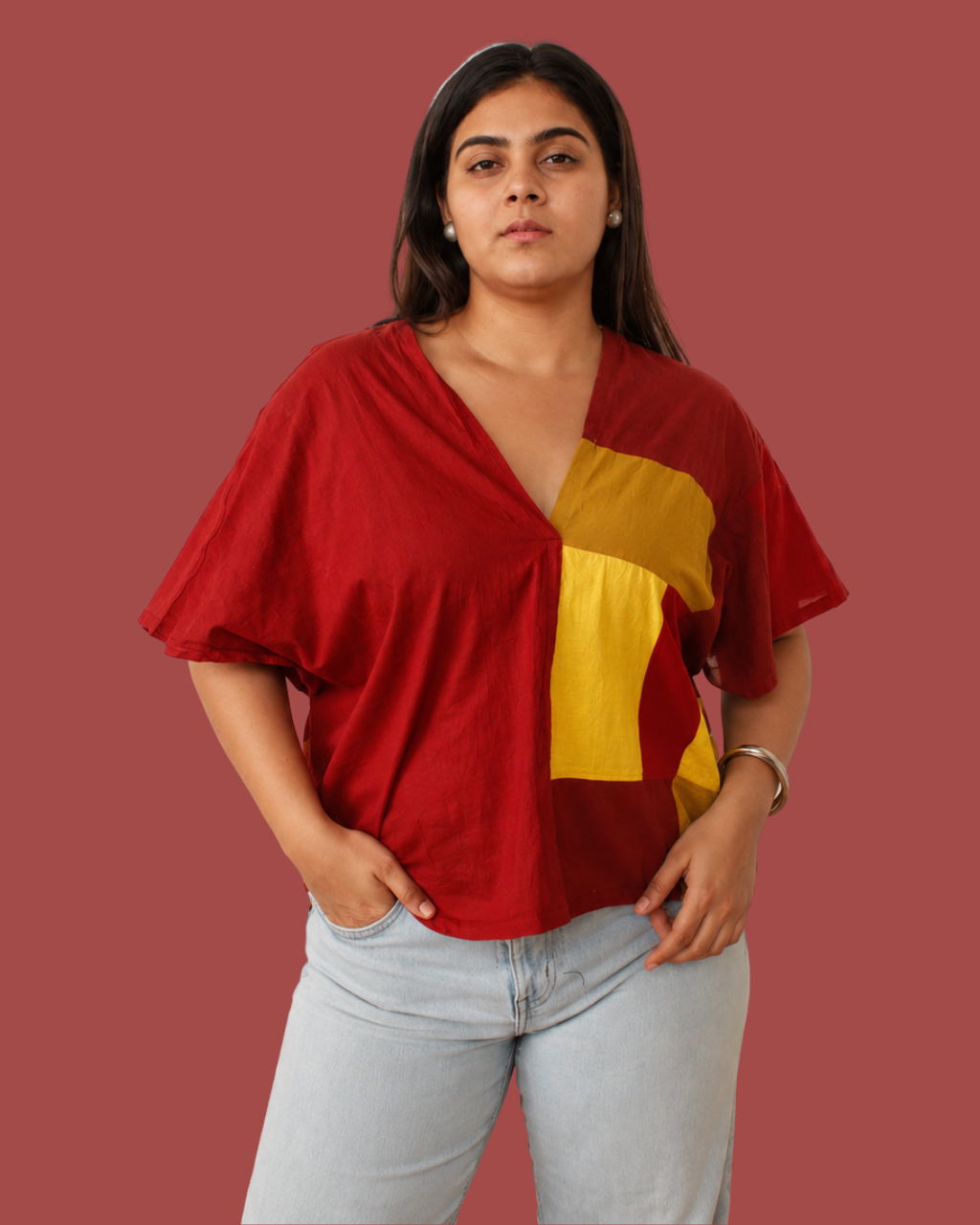 A woman modeling a red short-sleeve V-neck top with a large yellow geometric color-block panel on the front, paired with light-wash jeans, against a solid muted red background.