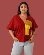 A woman modeling a red short-sleeve V-neck top with a large yellow geometric color-block panel on the front, paired with light-wash jeans, against a solid muted red background.