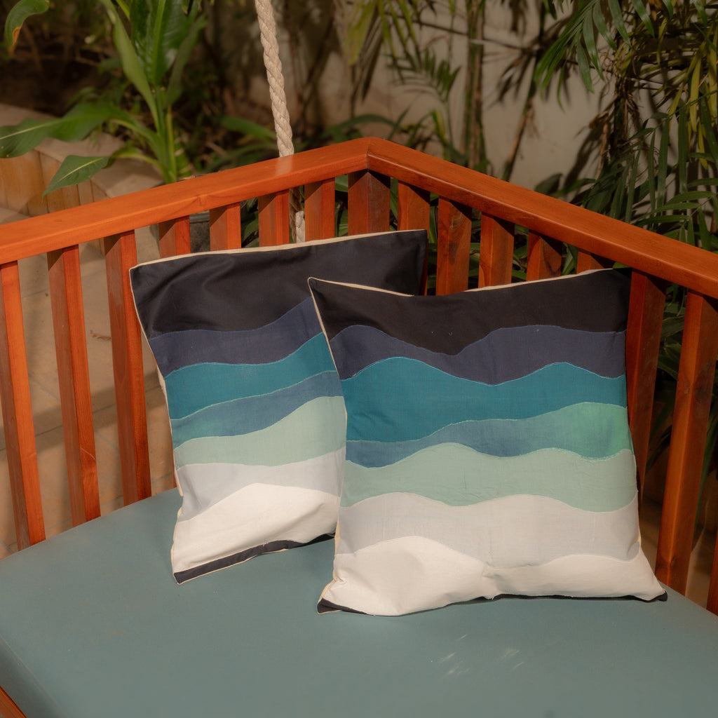 Two patterned pillows in shades of blue with a wooden railing and plants in the background