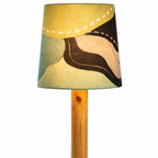 A table lamp with a cylindrical wooden base and stem, featuring a drum shade with an abstract painted pattern in olive green, beige, brown, and yellow tones. A black power cord extends from the base against a plain white background.