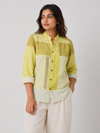 A woman wearing a green and cream patchwork button-up shirt with vertical leaf embroidery, paired with light beige trousers, against a plain light gray background.