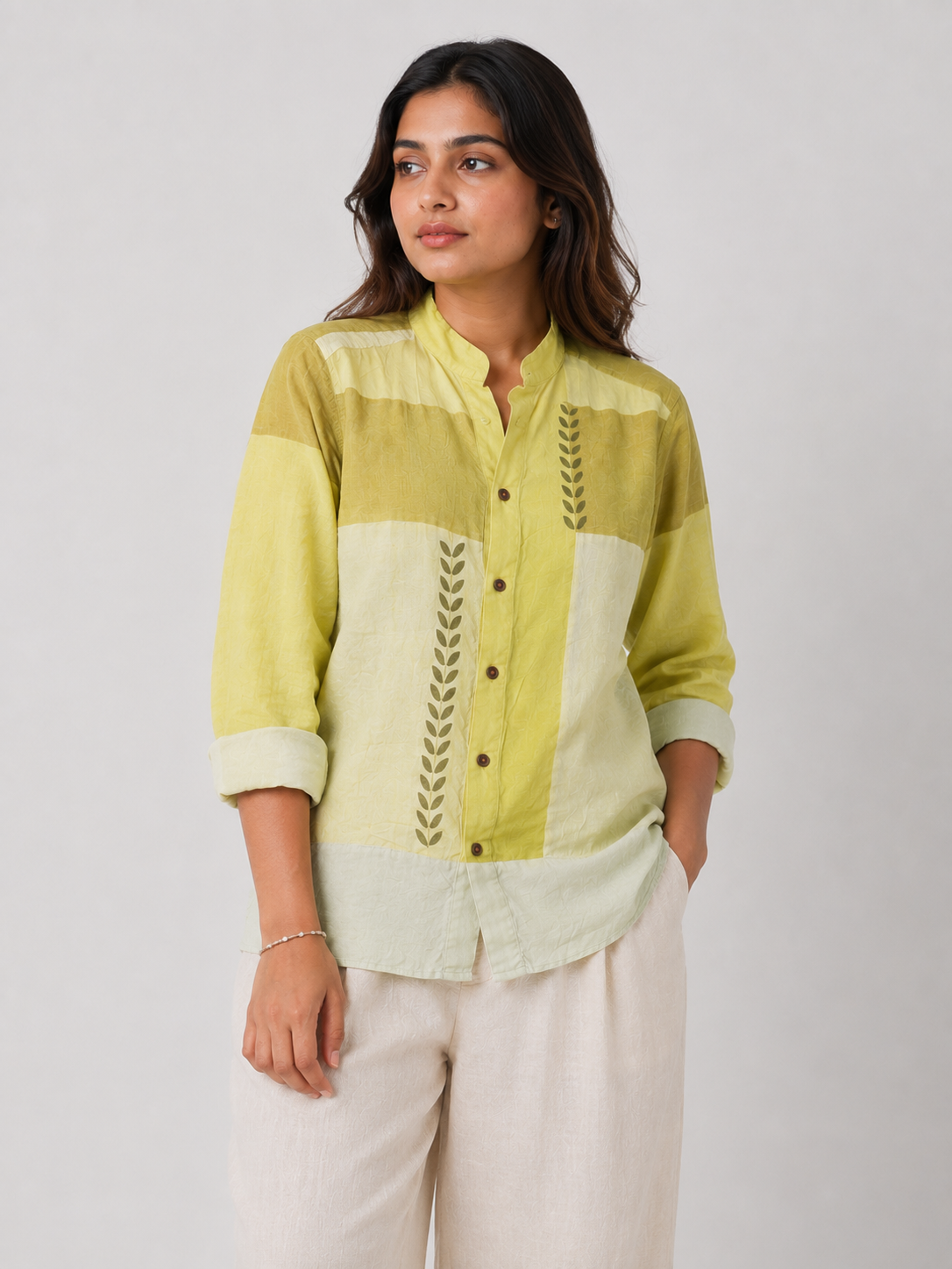 A woman wearing a green and cream patchwork button-up shirt with vertical leaf embroidery, paired with light beige trousers, against a plain light gray background.