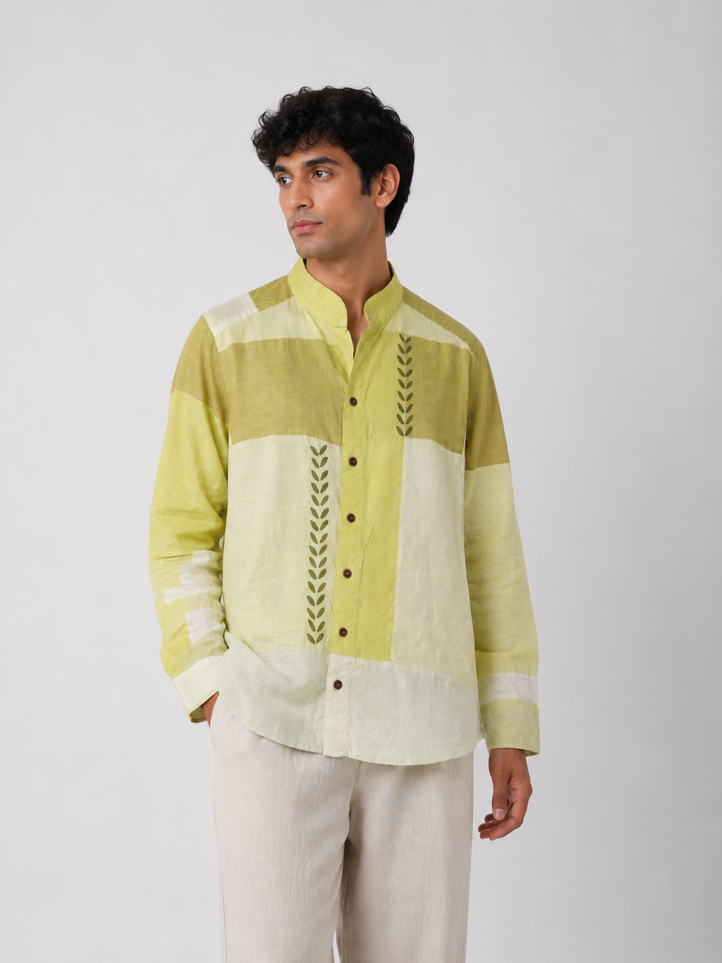 A man wearing a light green and off-white patchwork long-sleeve shirt with green leaf embroidery down the front, paired with off-white trousers, against a plain light gray background.