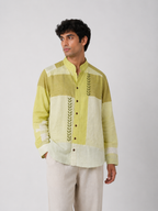 A man wearing a light green and off-white patchwork long-sleeve shirt with green leaf embroidery down the front, paired with off-white trousers, against a plain light gray background.