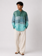 A man modeling a long-sleeve button-up shirt with horizontal stripes in shades of teal, blue, and mint green, paired with cream-colored trousers and brown sandals, against a light gray studio background.