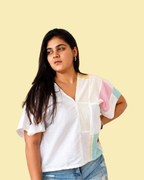 A woman wearing a white short-sleeve blouse with pastel pink, light blue, and yellow color-blocked panels on the sleeves and chest, paired with blue jeans, set against a solid pale yellow background.