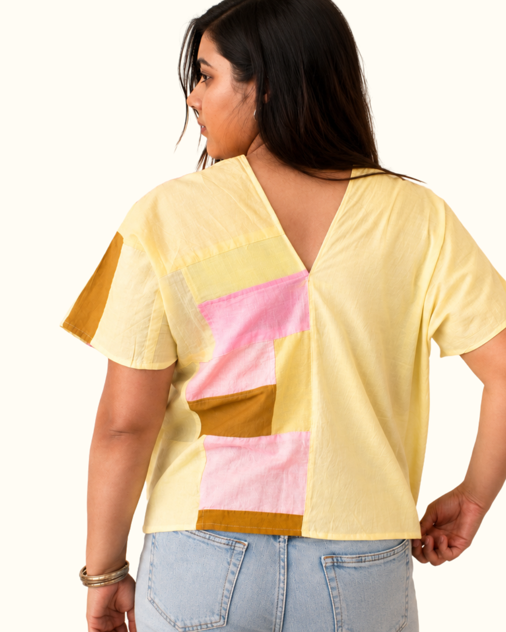 A woman shown from the back wearing a pale yellow short-sleeve top with a large patchwork panel in pink, yellow, and brown on the back, paired with light blue jeans against a plain white background.