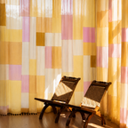 A set of sheer patchwork curtains in yellow, pink, and white hanging in front of a window, with two wooden lounge chairs in the foreground on a wooden floor.