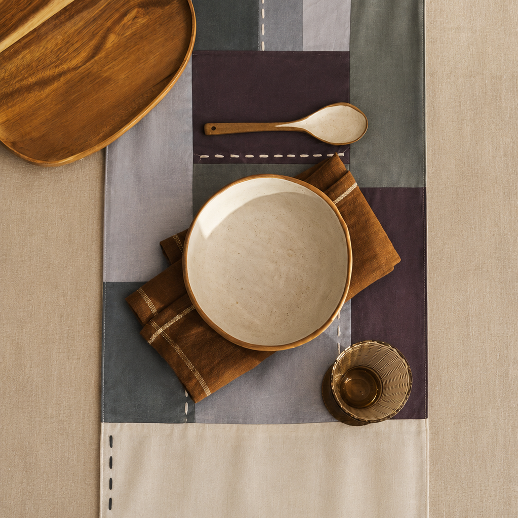 A styled flat-lay of a patchwork table runner with a cream lower section and stitched dashed borders, featuring blocks of gray, olive green, and dark purple. The runner is styled with a wooden plate, a ceramic bowl, a wooden spoon, and folded brown napkins on a beige linen tablecloth.