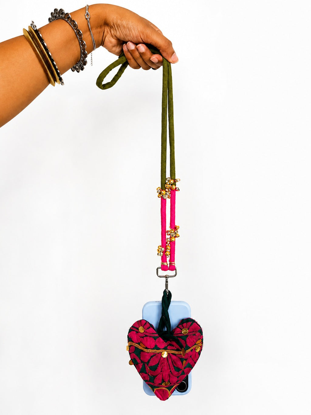 A hand wearing gold and silver bracelets holds a phone case with a long olive green strap, bright pink hanging cords, and a red and black patterned heart-shaped pouch with gold trim, against a plain white background.